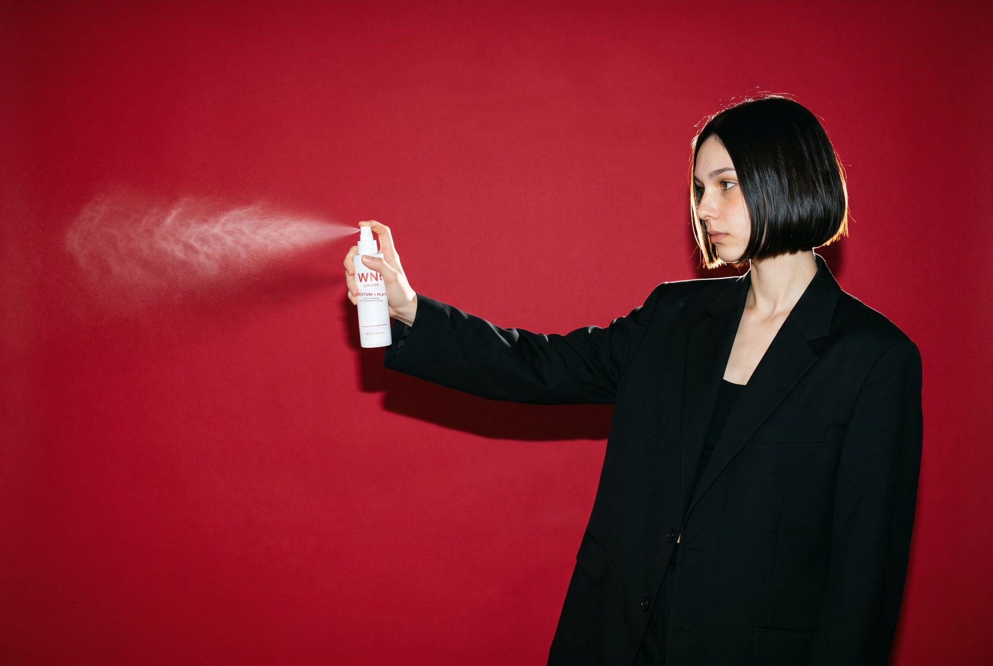 Model with a black bob in an oversized black suit spraying Restore + Play across a crimson wall