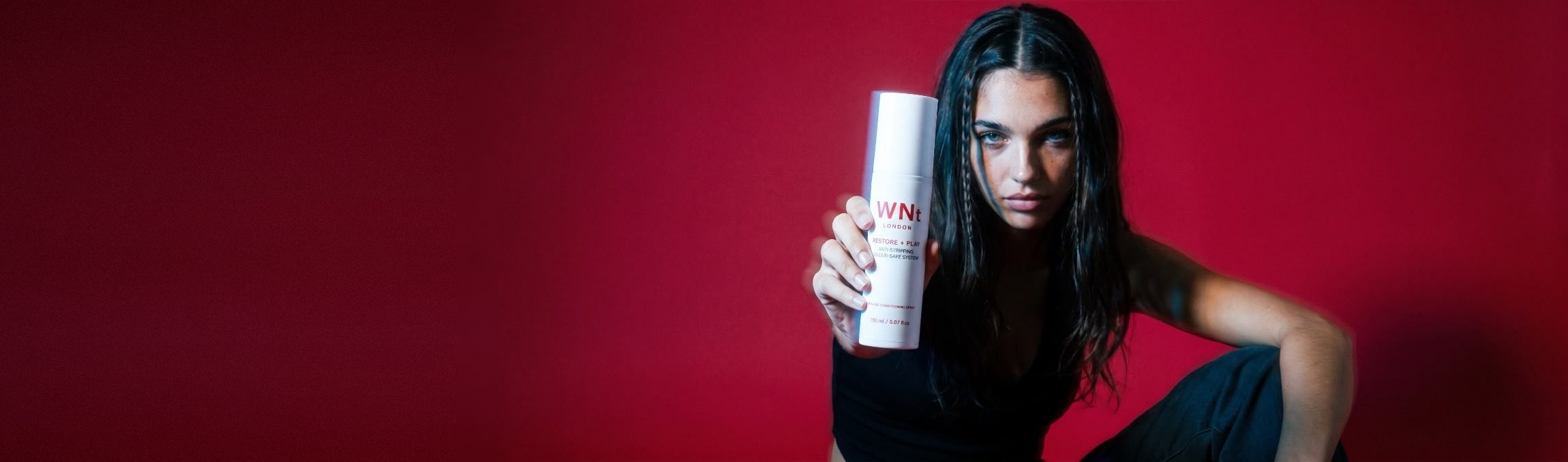 Model with long dark hair and fine braids, in black against a crimson wall, holding out WNt Restore + Play spray